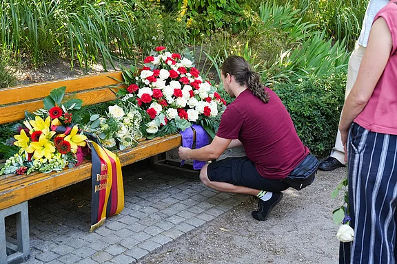 Ein Mann in einem roten T-Shirt kniet an einer Bank und legt einen Blumenstrauß ab. Neben ihm stehen mehrere Kränze aus roten und weißen Blumen sowie gelben Sonnenblumen. Im Hintergrund sind grüne Pflanzen zu sehen.