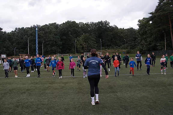 Auf einem Fußballplatz leitet eine Trainerin eine große Gruppe Kinder und Jugendlicher beim Aufwärmen. Die Teilnehmenden stehen verteilt in Reihen und folgen Bewegungsanweisungen, draußen bei bewölktem Himmel.