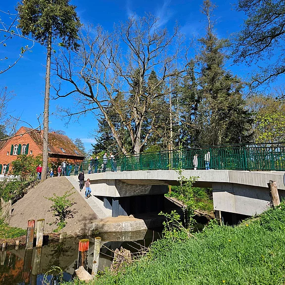 Fußgängerbrücke mit grünem Geländer über einen kleinen Bach in einem Park. Einige Menschen gehen darüber, eine Person steigt die Treppe hinunter zum Ufer. Frühlingsbäume und blauer Himmel; rotes Backsteinhaus im Hintergrund.