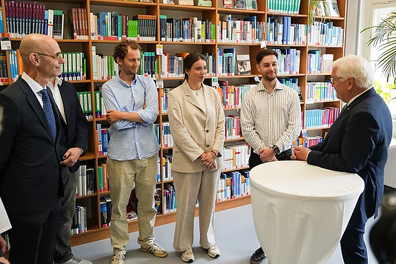 Eine Gruppe von fünf Personen steht in einer Bibliothek vor einem Regal mit Bücher. Eine Person spricht mit einer anderen, während die anderen aufmerksam zuhören. Alle sind in formeller Kleidung. Im Vordergrund steht ein runder Tisch.