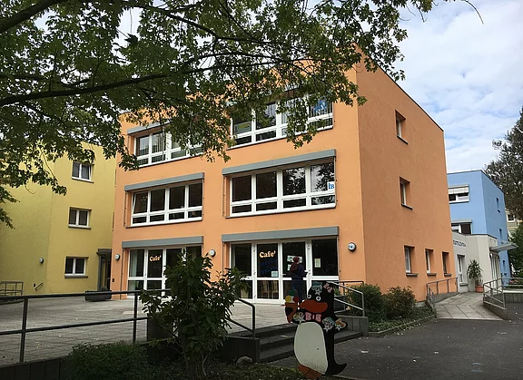 Ein dreistöckiges, orangefarbenes Gebäude mit großen Fenstern, das ein Café beherbergt. Vor dem Eingang steht eine saisonale Deko in Form eines Pinguins, der ein Eis hält. Im Hintergrund sind weitere bunte Gebäude zu sehen.