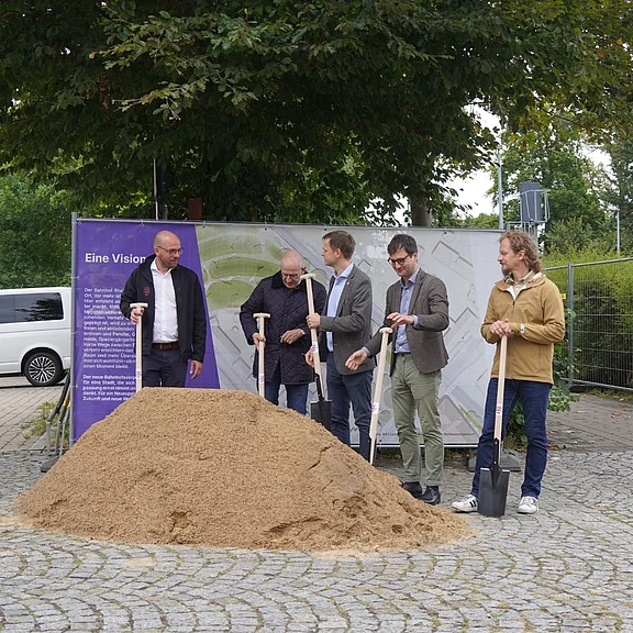 Fünf Personen halten Spaten vor einem Sandhaufen und posieren beim feierlichen Spatenstich eines Bauvorhabens.