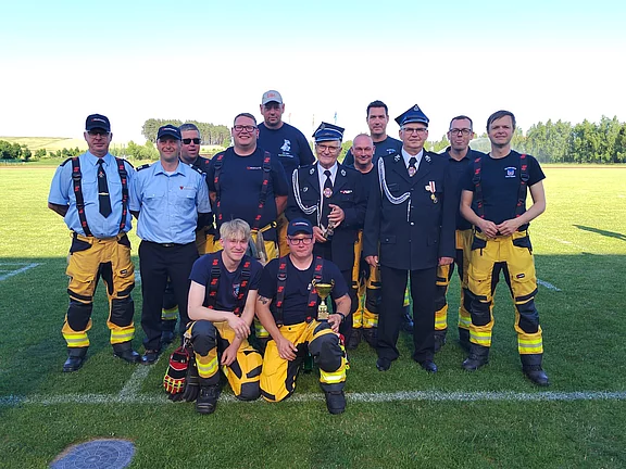 Gruppenfoto von Feuerwehrleuten im Freien: mehrere Personen in gelben Einsatzhosen und schwarzen T‑Shirts, zwei in dunklen Ausgehuniformen mit Mützen und Abzeichen. Sie stehen und knien auf einer Rasenfläche bei Tageslicht und blicken in die Kamera.
