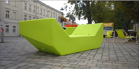 Eine große, leuchtend grüne Sitzgelegenheit in moderner, geometrischer Form steht auf einem Pflastersteinboden. Im Hintergrund sind einige Stühle und Gebäude sichtbar, die eine einladende Atmosphäre schaffen.