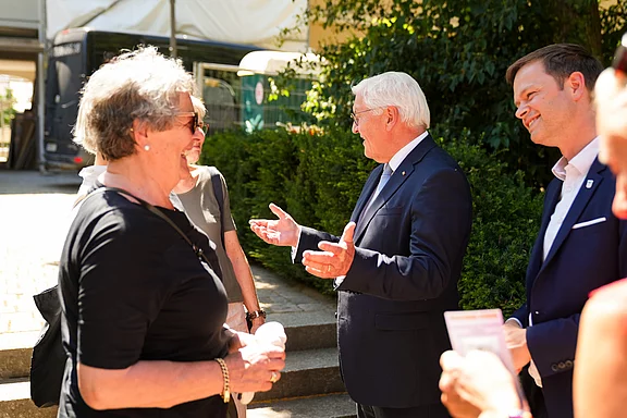 Eine Gruppe von Menschen steht im Freien und unterhält sich. Ein älterer Mann in Anzug spricht mit einer Frau, die lächelt. Weitere Personen hören zu und lachen. Die Szene vermittelt eine freundliche, gesellige Atmosphäre.