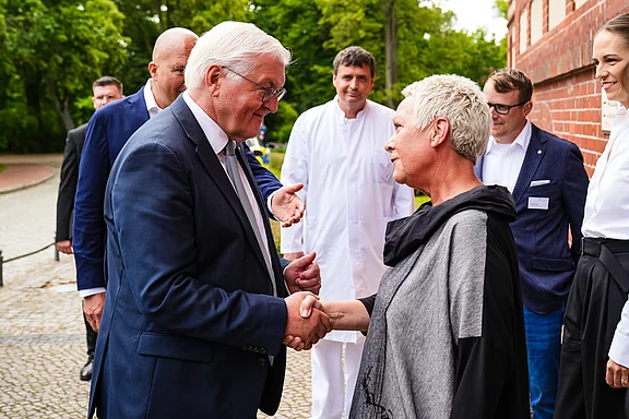 Ein Mann im Anzug schüttelt einer Frau mit kurzem, grauem Haar die Hand. Im Hintergrund sind weitere Personen sichtbar, die den Austausch beobachten. Die Szene findet in einem grünen Umfeld statt, umgeben von Bäumen und einer roten Backsteinwand.
