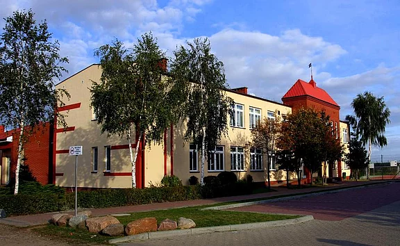Zweigeschossiges, hellgelbes Gebäude mit roten Akzenten und turmartigem Eckteil. Große Fenster entlang der Fassade. Birken säumen einen gepflasterten Gehweg. Im Vordergrund eine ruhige Straße, darüber blauer Himmel mit Wolken.