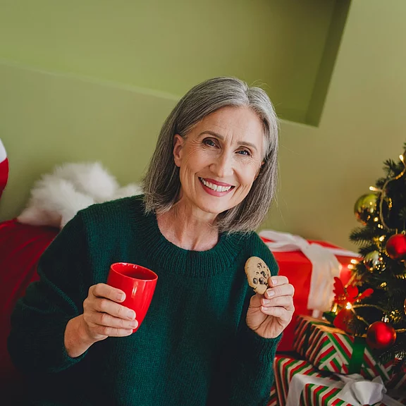 Ältere Frau mit grauen Haaren sitzt lächelnd neben einem geschmückten Weihnachtsbaum. Sie trägt einen dunkelgrünen Pullover, hält eine rote Tasse und einen Keks. Geschenkpakete im Hintergrund, warme, festliche Stimmung.