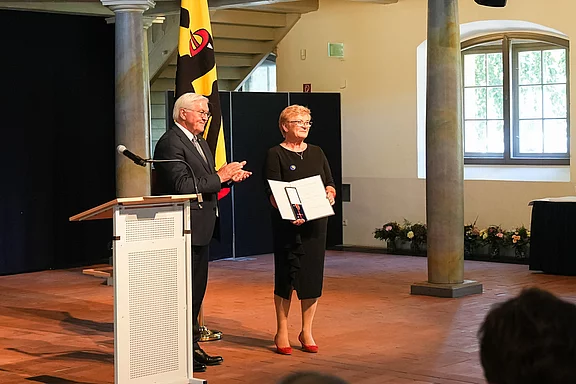 Eine Frau in schwarzem Kleid erhält eine Auszeichnung von einem Mann in Anzug. Beide stehen auf einer Bühne mit einem Rednerpult im Vordergrund. Im Hintergrund sind Pflanzen zu sehen und eine Fahne, die auf das Ereignis hinweist.