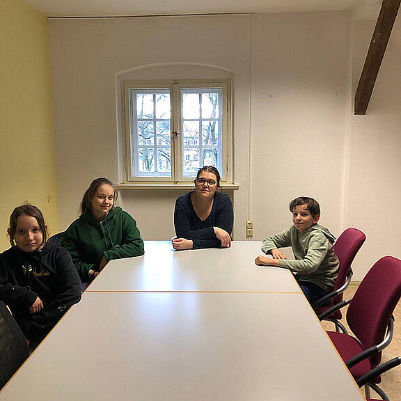 Vier Kinder und eine junge Frau sitzen an einem Tisch in einem hellen Raum. Sie blicken in die Kamera, die Wand ist gelb gestrichen und ein Fenster mit Blick auf Bäume ist geöffnet. Sie wirken entspannt und aufmerksam.