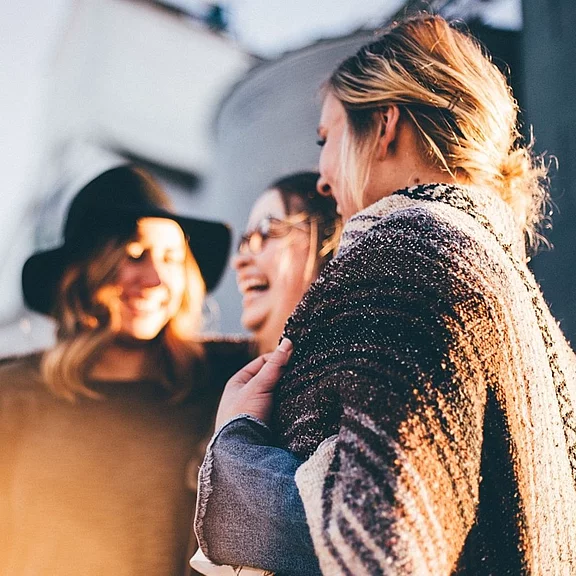 Drei junge Erwachsene stehen draußen dicht beieinander, lachen und unterhalten sich. Warmes Licht schafft eine gemütliche Stimmung; eine Person trägt einen Hut, eine andere ist in eine Decke gehüllt. Der Moment vermittelt Freundschaft und Geselligkeit.
