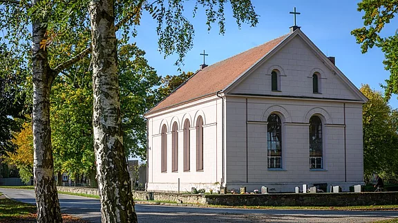 Ein kleines, helles Gebäude mit einem Satteldach und typischen Kirchenkreuz auf dem Dach. Die Fassade hat große Bogenfenster und ist von Bäumen und einem ruhigen Weg umgeben. Der Platz vor der Kirche ist mit Blumenschmuck dekoriert.