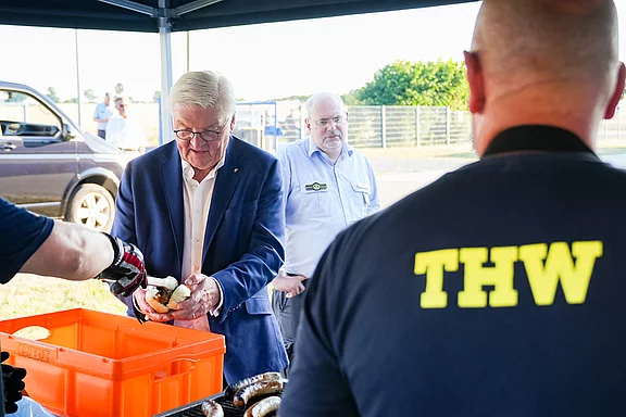 Ein älterer Mann mit Brille und Anzug hält eine Wurst in der Hand, während er an einem Stand steht, wo Speisen zubereitet werden. Im Hintergrund sind weitere Personen sichtbar, darunter einer mit einem T-Shirt mit der Aufschrift „THW“.