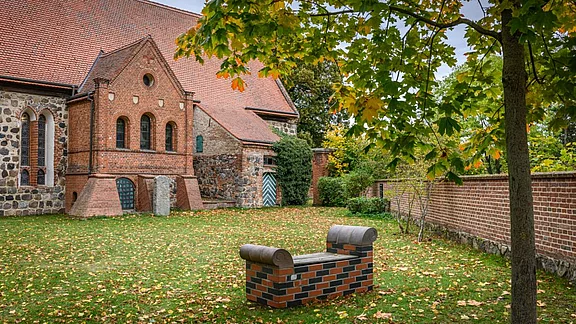 Eine alte Backsteinkirche steht im Hintergrund, umgeben von einer grünen Wiese mit herabgefallenen Blättern. Im Vordergrund befindet sich eine ungewöhnliche Bank, die wie ein Mauerwerk aussieht. Darüber ist die Baumkrone, die teilweise gelbe Blätter zeigt.