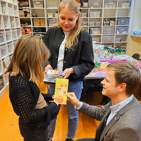 In einem Bastelraum zeigt ein Kind einem sitzenden Mann eine selbstgebastelte, bunte Karte mit Blumenmotiv. Eine Frau daneben hält Malutensilien und beobachtet lächelnd. Der Fokus liegt auf der gemeinsamen Kreativarbeit und dem Präsentieren des Ergebnisses.