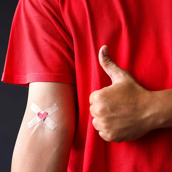 Person im roten T‑Shirt zeigt Daumen hoch. Am Oberarm klebt ein gekreuztes Pflaster mit kleinem Herz, wie nach einer Blutspende oder Impfung. Positive, unterstützende Botschaft zur Gesundheit und Hilfsbereitschaft.
