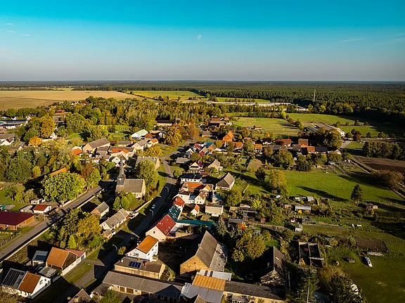 Eine Luftaufnahme eines kleinen Dorfes, umgeben von Wäldern und Feldern. Die Wohnhäuser sind bunt, mit Gärten und Straßen, die durch die Landschaft führen. Die Umgebung zeigt eine ruhige, ländliche Atmosphäre unter einem blauen Himmel.