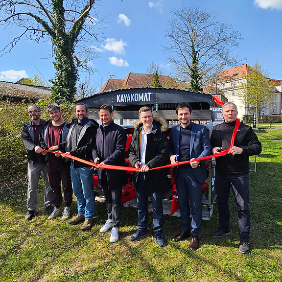 Acht Personen stehen im Freien vor einem KAYAKOMAT-Stand und halten gemeinsam ein langes rotes Band als Zeichen einer Eröffnung. Hinter ihnen sind rote Kajaks auf einem Anhänger zu sehen.