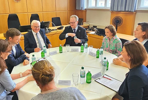 Eine Gruppe von sechs Personen sitzt an einem runden Tisch, umgeben von Wasserflaschen. Sie diskutieren miteinander, während eine Person etwas zu erklären scheint. Der Raum hat eine formelle Atmosphäre, mit Holzverkleidung und Fenstern im Hintergrund.