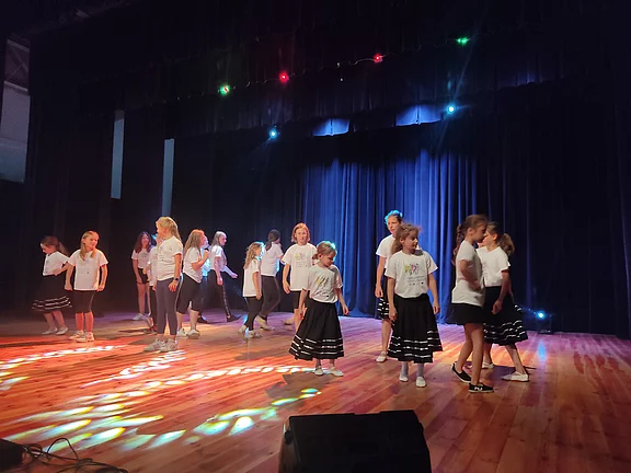 Eine Gruppe Kinder und Jugendlicher tanzt auf einer Theaterbühne. Sie tragen weiße T‑Shirts; einige Mädchen schwarze Röcke. Bunte Scheinwerfer beleuchten den Holzboden, hinter ihnen ein dunkelblauer Vorhang. Gruppenchoreografie in Aufführung.