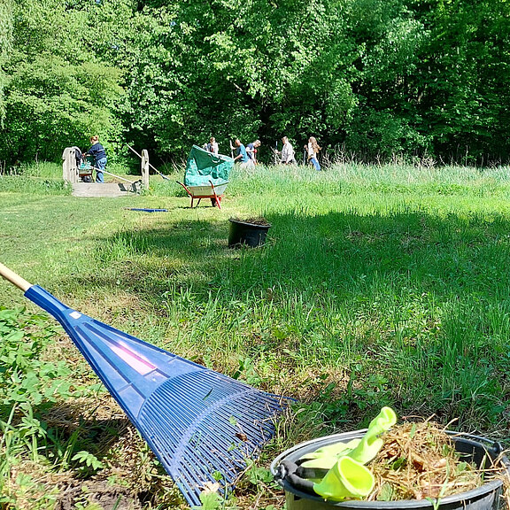 Eine grüne Wiese mit einer blauen Harke im Vordergrund. Im Hintergrund sind mehrere Personen zu sehen, die sich in einer ruhigen Umgebung aufhalten. Bäume umgeben den Bereich, und im Hintergrund ist eine kleine Holzplattform zu erkennen.