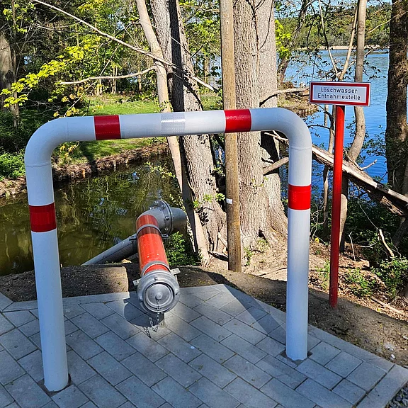 Feuerwehr-Wasserentnahmestelle am Ufer: Ein rot-weiß markiertes Schutzgestell umgibt einen Anschlussstutzen mit Rohr zur Wasserentnahme. Daneben weist ein Schild mit der Aufschrift „Löschwasser Entnahmestelle“ darauf hin.