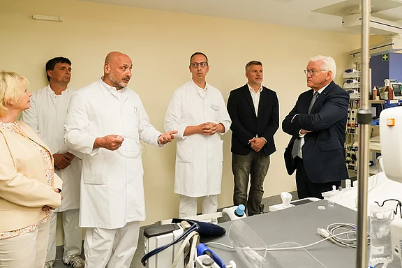 Eine Gruppe von fünf Personen steht in einem medizinischen Umfeld. Ein männlicher Arzt erklärt etwas, während die anderen zuhören. Unter den Zuhörern sind auch Personen in formeller Kleidung, die anscheinend an der Diskussion teilnehmen. Im Hintergrund sind medizinische Geräte sichtbar.