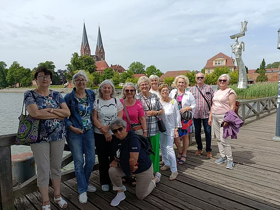 Eine Gruppe von zwölf älteren Frauen steht auf einem Holzsteg neben einem Gewässer. Im Hintergrund sind Bäume und zwei markante Kirchtürme sichtbar. Die Frauen lächeln und tragen unterschiedliche, sommerliche Kleidung.