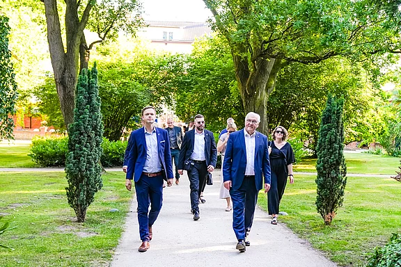 Eine Gruppe von sieben Personen in Anzügen und eleganter Kleidung geht auf einem Weg durch einen parkähnlichen Garten, umgeben von Bäumen und Grünflächen. Die Atmosphäre ist freundlich und entspannt.