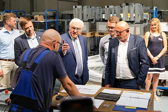 Eine Gruppe von sechs Personen steht um einen Tisch in einer Werkstatt. Eine Person im Blaumann erklärt etwas auf einem Papier. Der Fokus liegt auf der Interaktion zwischen den Personen, die interessiert zuhören, während ein weiterer Mann im Anzug aufmerksam schaut.