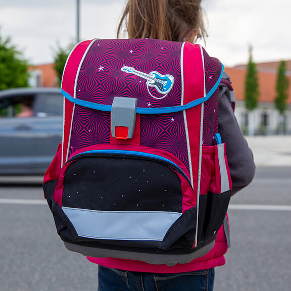 Ein Mädchen trägt einen bunten Rucksack in Pink und Schwarz, dekoriert mit einem Gitarrenmotiv. Der Rucksack hat eine auffällige Form und eine praktische Verschlussmechanik. Im Hintergrund sind verschwommen Bäume und ein vorbeifahrendes Auto zu sehen.