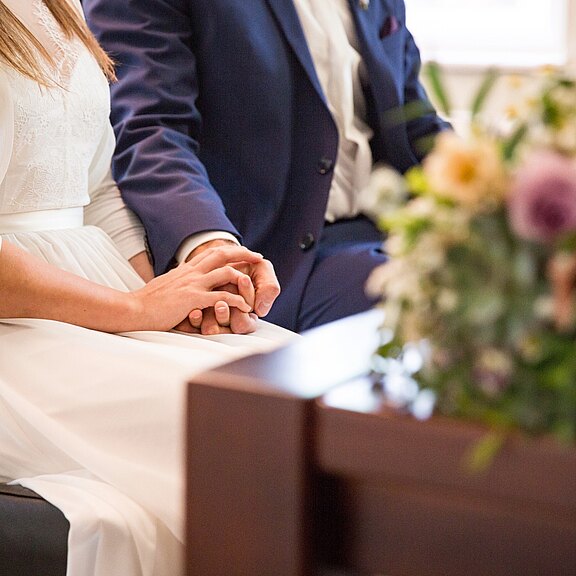 Ein Brautpaar sitzt nebeneinander und hält sich liebevoll an den Händen. Die Frau trägt ein weißes Kleid, der Mann einen blauen Anzug. Weicher, heller Hintergrund; rechts unscharfe Blumen. Fokus liegt auf den verbundenen Händen und der ruhigen, feierlichen Stimmung.