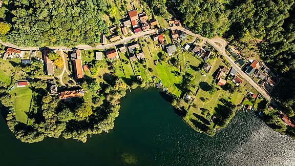Luftaufnahme eines kleinen Dorfes am Wasser, umgeben von Bäumen. Die Häuser sind in einer Linie entlang einer Straße angeordnet, mit grünen Wiesen und Gärten. Der See reflektiert Sonnenlicht und ist von Pflanzen und Booten gesäumt.