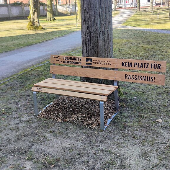 Holzbank im Park mit absichtlich halbiertem Sitz. Auf der Rückenlehne steht groß: „Kein Platz für Rassismus!“. Logos von „Tolerantes Brandenburg“ und „Fontanestadt Neuruppin“. Die Bank symbolisiert, dass Ausgrenzung keinen Raum hat.
