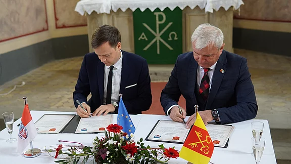 Zwei Anzugträger sitzen an einem Tisch und unterschreiben offizielle Dokumente. Vor ihnen stehen kleine Tischflaggen, darunter die EU-Flagge, sowie Blumen. Die Szene wirkt wie eine formelle Unterzeichnung oder Partnerschaftsvereinbarung in einem feierlichen Raum.