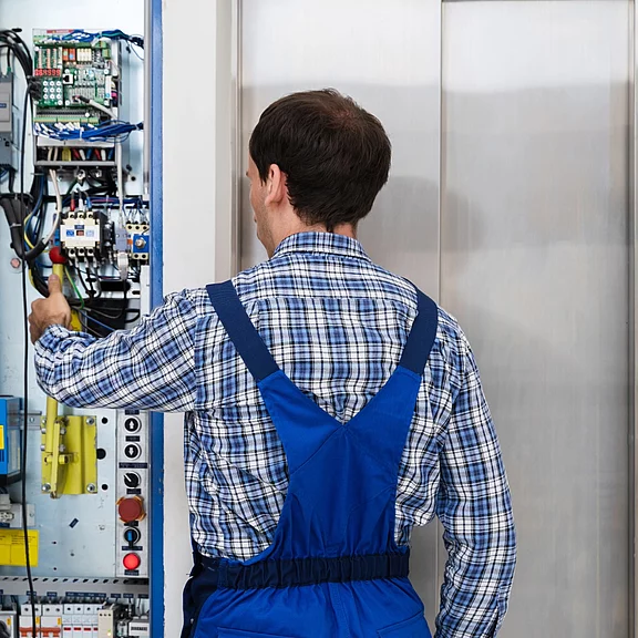 Techniker in blauer Arbeitskleidung steht vor offenem Schaltschrank und justiert mit einem Schraubendreher die Elektronik einer Aufzugsanlage neben geschlossenen Aufzugstüren.