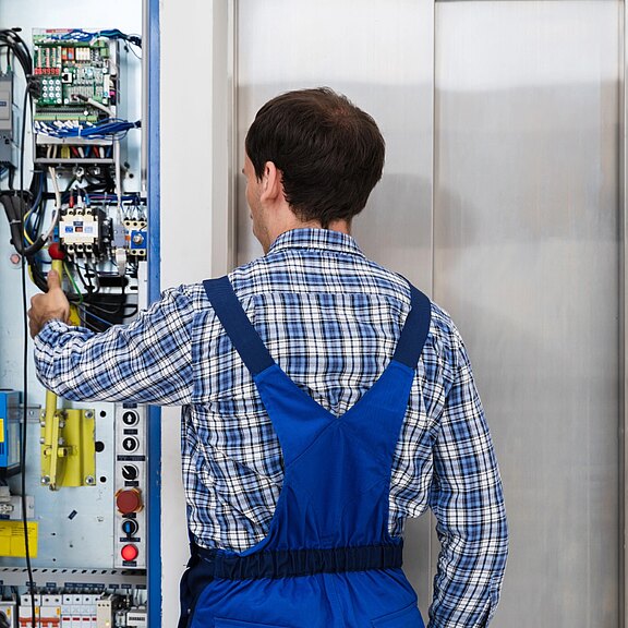 Techniker in blauer Arbeitskleidung steht vor offenem Schaltschrank und justiert mit einem Schraubendreher die Elektronik einer Aufzugsanlage neben geschlossenen Aufzugstüren.