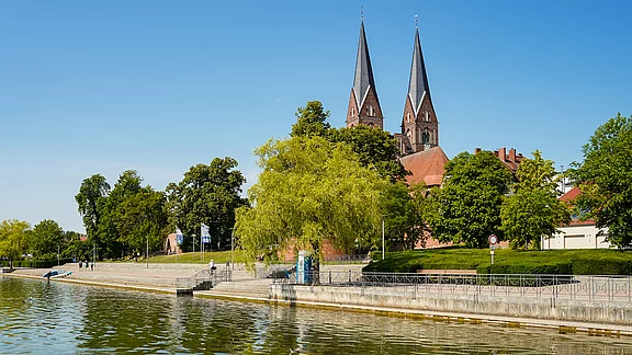 Ein ruhiger Uferbereich mit Bäumen und einer Stufe, die zum Wasser führt. Im Hintergrund sind zwei hohe Türme einer Kirche sichtbar. Der Himmel ist klar und blau, und einige Spaziergänger sind in der Nähe zu sehen.
