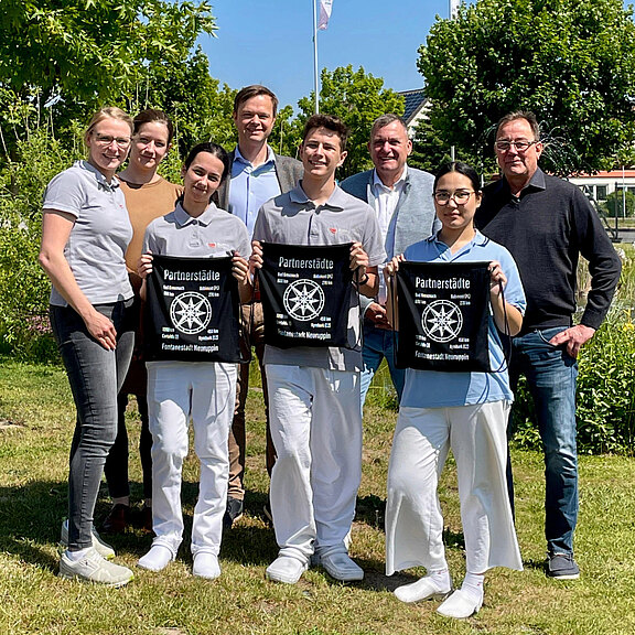 Eine Gruppe von sieben Personen steht im Freien und lächelt in die Kamera. Zwei Personen in Uniform halten T-Shirts mit dem Aufdruck „Partnerstadt – Gemeinschaft gestalten“. Der Hintergrund zeigt grüne Pflanzen und einen klaren Himmel.