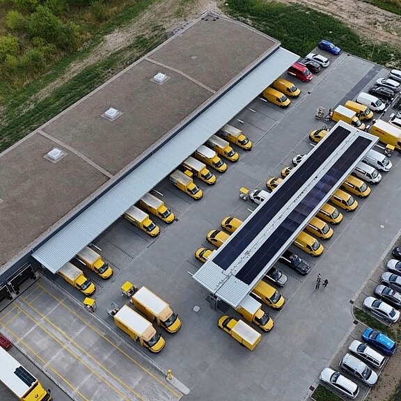 Luftaufnahme eines Logistikzentrums: Dutzende gelbe Lieferwagen stehen in geordneten Reihen neben einer Halle und unter einem langen überdachten Bereich mit Solarpanelen, offenbar zum Laden oder Bereitstellen für Touren. Markierte Ladezonen und Einfahrten strukturieren den Hof.