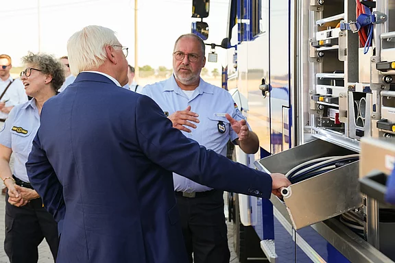 Eine Gruppe von Personen steht vor einem Feuerwehrfahrzeug. Ein Mann im Anzug zeigt auf den Geräteschrank des Fahrzeugs, während ein Feuerwehrmann ihm etwas erklärt. Eine weitere Person hört aufmerksam zu. Die Szene deutet auf eine Informationsveranstaltung zur Feuerwehrausrüstung hin.