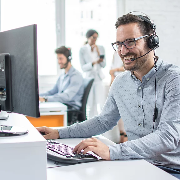 Freundlicher Kundenservice-Mitarbeiter mit Headset arbeitet lächelnd am Computer in einem hellen Büro; im Hintergrund arbeiten weitere Kollegen. Symbolisiert telefonischen Support, Teamarbeit und professionelle Betreuung.