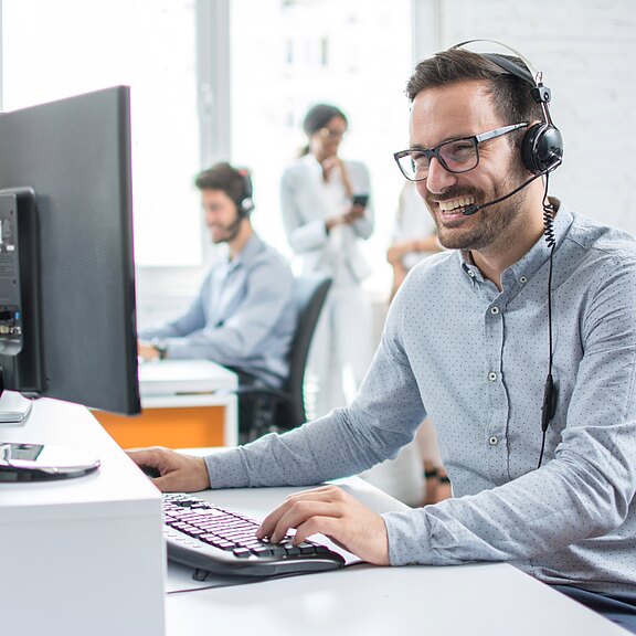 Freundlicher Kundenservice-Mitarbeiter mit Headset arbeitet lächelnd am Computer in einem hellen Büro; im Hintergrund arbeiten weitere Kollegen. Symbolisiert telefonischen Support, Teamarbeit und professionelle Betreuung.