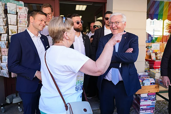 Eine Gruppe von Menschen steht vor einem Geschäft. Eine Frau spricht mit einem älteren Mann, der lächelt. Die Umgebung zeigt Bücher und Karten. Die Stimmung wirkt freundlich und offen, mit einigen Personen im Hintergrund, die aufmerksam zuhören.