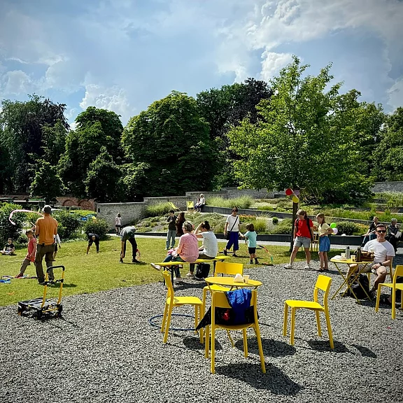 Lebhafter Park: Kinder spielen auf einer Wiese, Erwachsene sitzen und unterhalten sich. Gelbe Stühle und ein Tisch im Vordergrund, dahinter ein Teich und viele Bäume. Sommerliche, entspannte Atmosphäre.