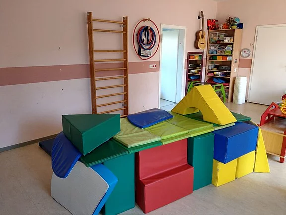 Der Raum zeigt eine Spielumgebung mit bunten Schaumstoffbausteinen, die zu einer Struktur aufgebaut sind. Im Hintergrund befindet sich ein Klettergerüst. Es handelt sich um eine kinderfreundliche Umgebung, die zum Spielen und Entdecken anregt.