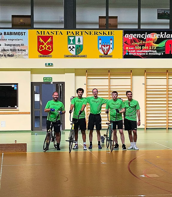 Fünf Radfahrer stehen in einem Sportzentrum, tragen grüne Teamshirts und halten ihre Fahrräder. Der Raum hat Holzfußboden und an der Wand sind verschiedene farbige Banner angebracht. Die Athleten wirken freundlich und stolz auf ihre Teamzugehörigkeit.