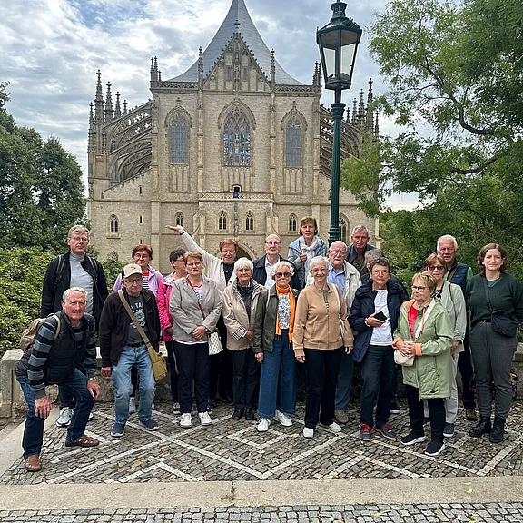 Gruppe von rund zwanzig Erwachsenen, überwiegend älteren Reisenden, posiert lächelnd auf einem gepflasterten Platz vor einer großen gotischen Kirche mit spitzen Türmen und filigranen Strebewerken; Bäume rahmen die Szene.