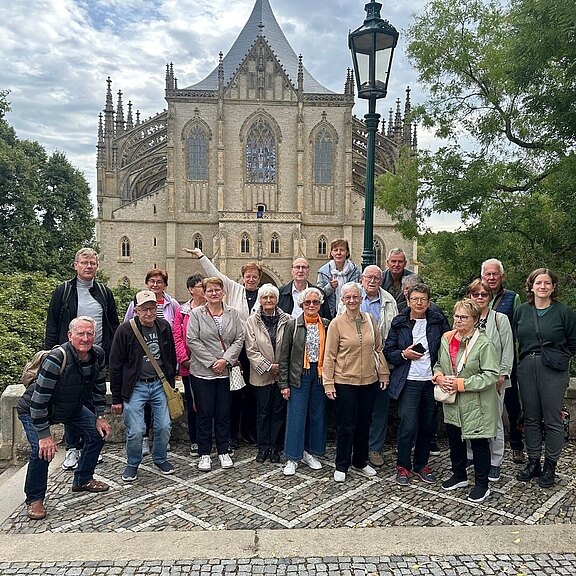 Gruppe von rund zwanzig Erwachsenen, überwiegend älteren Reisenden, posiert lächelnd auf einem gepflasterten Platz vor einer großen gotischen Kirche mit spitzen Türmen und filigranen Strebewerken; Bäume rahmen die Szene.