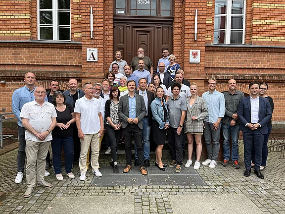 Gruppenfoto vor einem Backsteingebäude: Rund drei Dutzend Menschen stehen und sitzen auf Treppenstufen und lächeln in die Kamera. Mischung aus Männern und Frauen, leger bis leicht formell gekleidet. Hintergrund: Eingangstür und Fenster des Gebäudes.